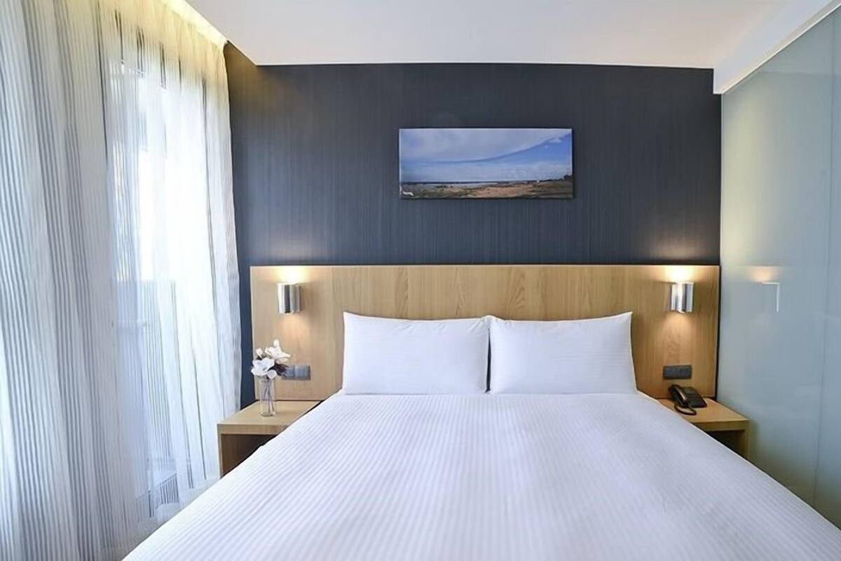 A hotel room with a large bed, two bedside tables with lamps, a painting on the wall, and a view of the outdoors.