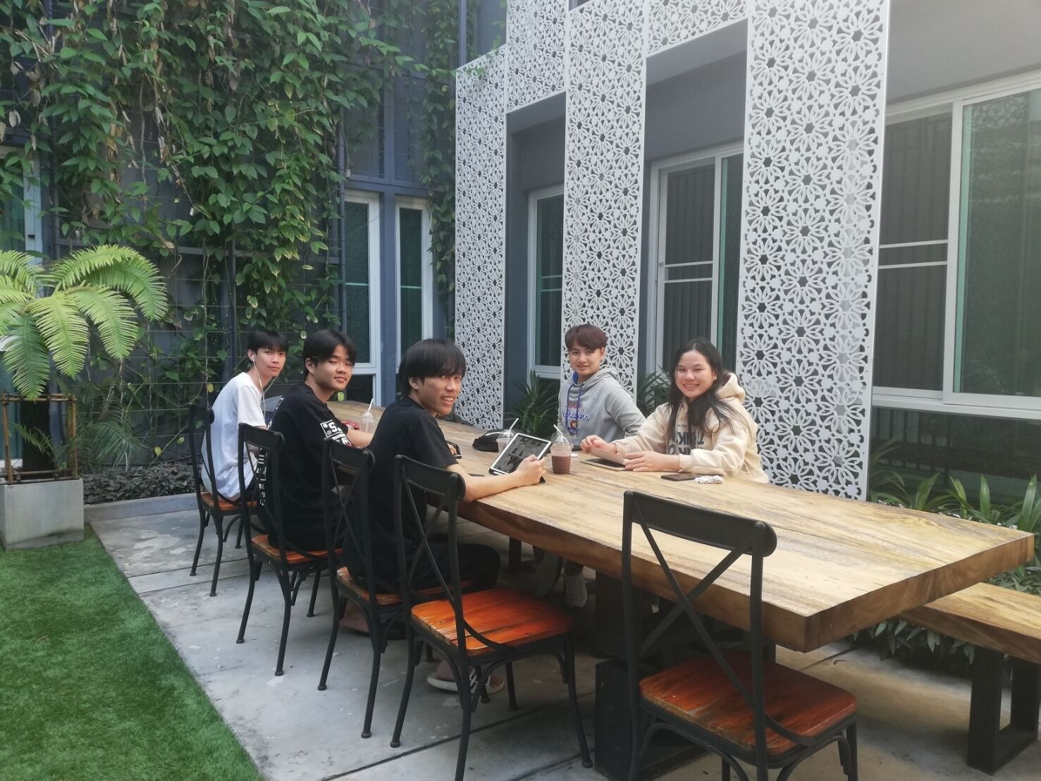 A group of people seated around a wooden table in an outdoor setting with a modern building in the background.