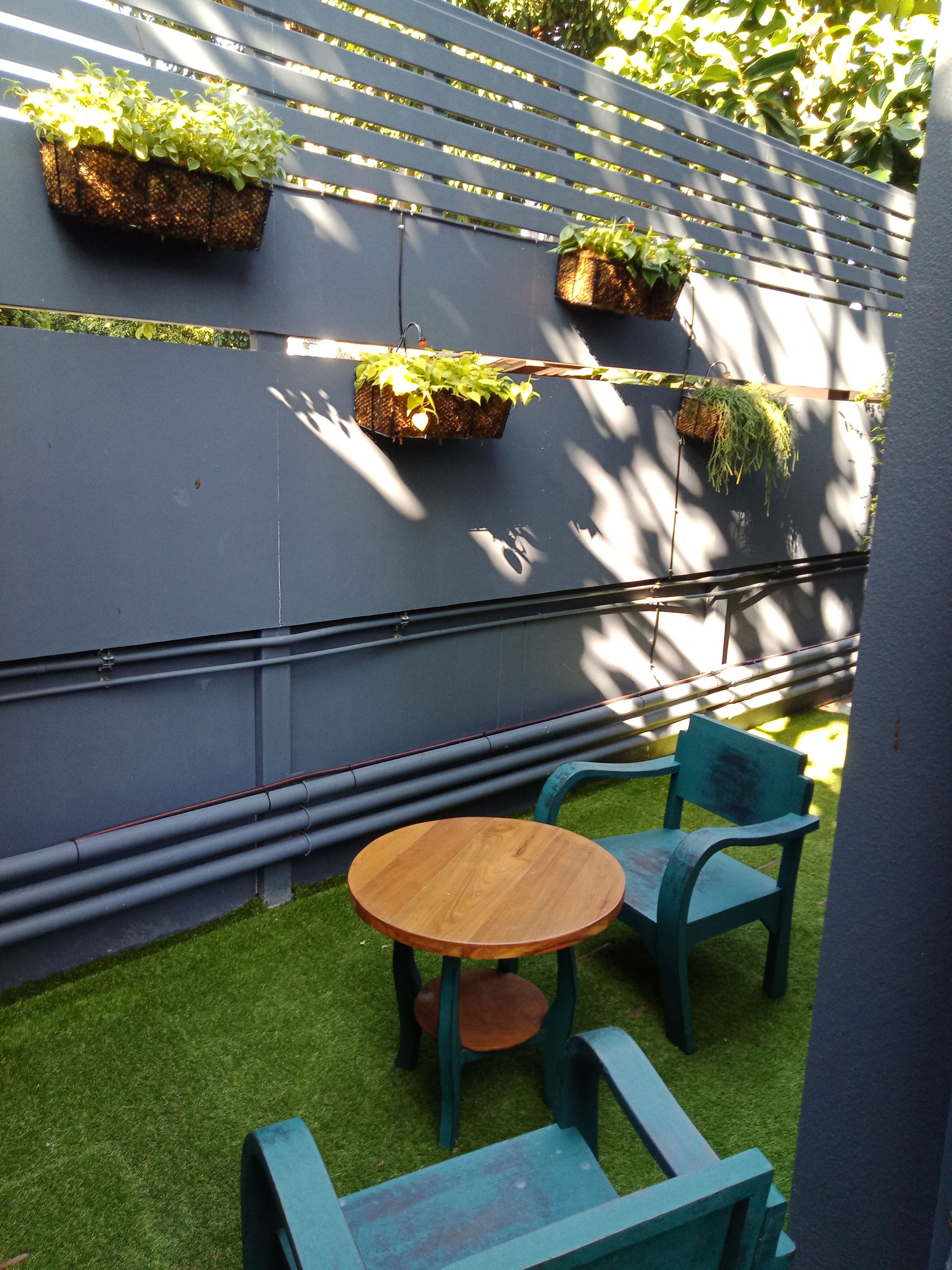 A patio with a round wooden table and two green chairs, a grey wall with hanging planters, and a small patch of artificial grass.