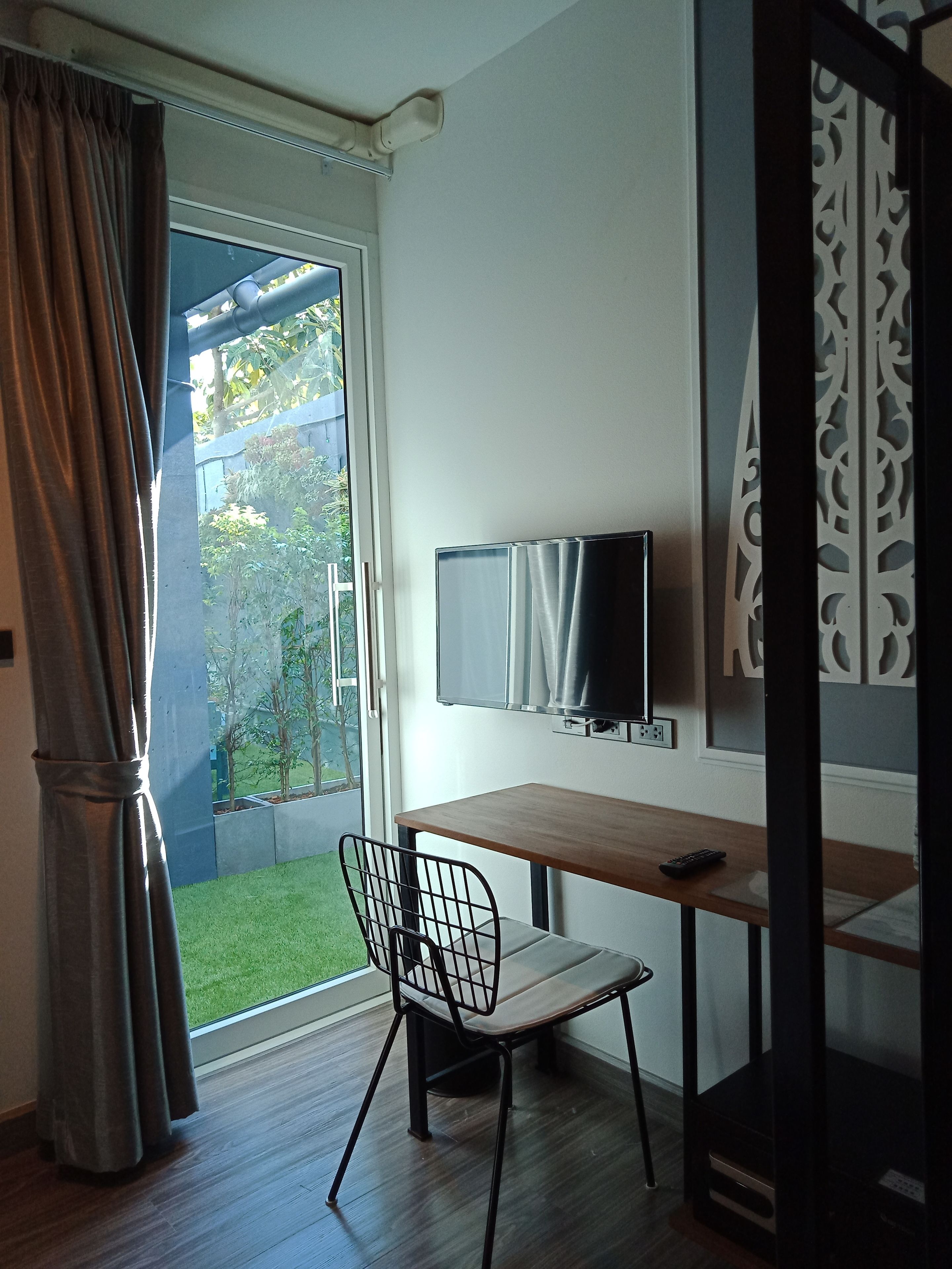 Una habitación con un escritorio de madera, una silla, un televisor y vistas a un jardín a través de un ventanal.
