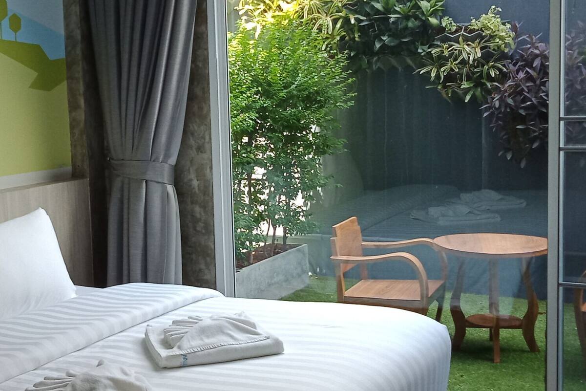 A hotel room with a bed, a small table, and chairs, a balcony with plants, and a view of greenery.