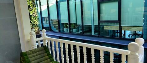 A white bench with a green cushion on a balcony with white balusters, overlooking a modern building with large windows.