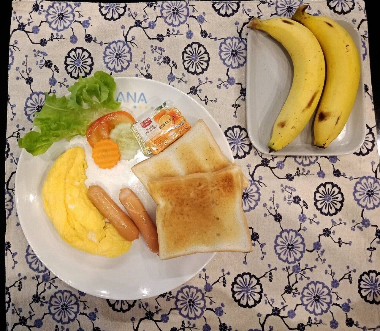 A plate with scrambled eggs, a hot dog, toast, lettuce, tomatoes, and a banana.