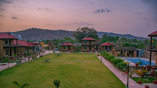 A courtyard with a swimming pool, surrounded by buildings with red roofs and a hilly landscape in the background.