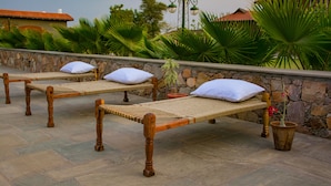 Two wooden beds with woven mattresses and white pillows, placed on a stone-paved terrace with a stone wall and lush green plants in the background.
