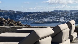 Vista para o mar, com uma fileira de cadeiras de descanso bege em deck de madeira, com vista para uma cidade costeira com edifícios brancos e céu claro.