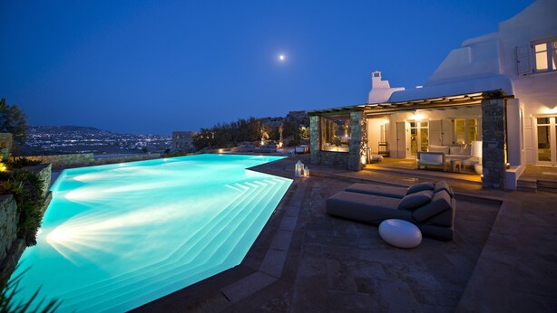 Uma villa moderna com piscina, arquitetura em pedra e vista da cidade ao entardecer.