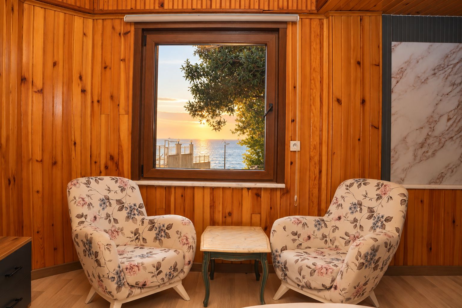 Une pièce aux murs lambrissés, une fenêtre donnant sur la mer, deux fauteuils à motifs floraux et une petite table.