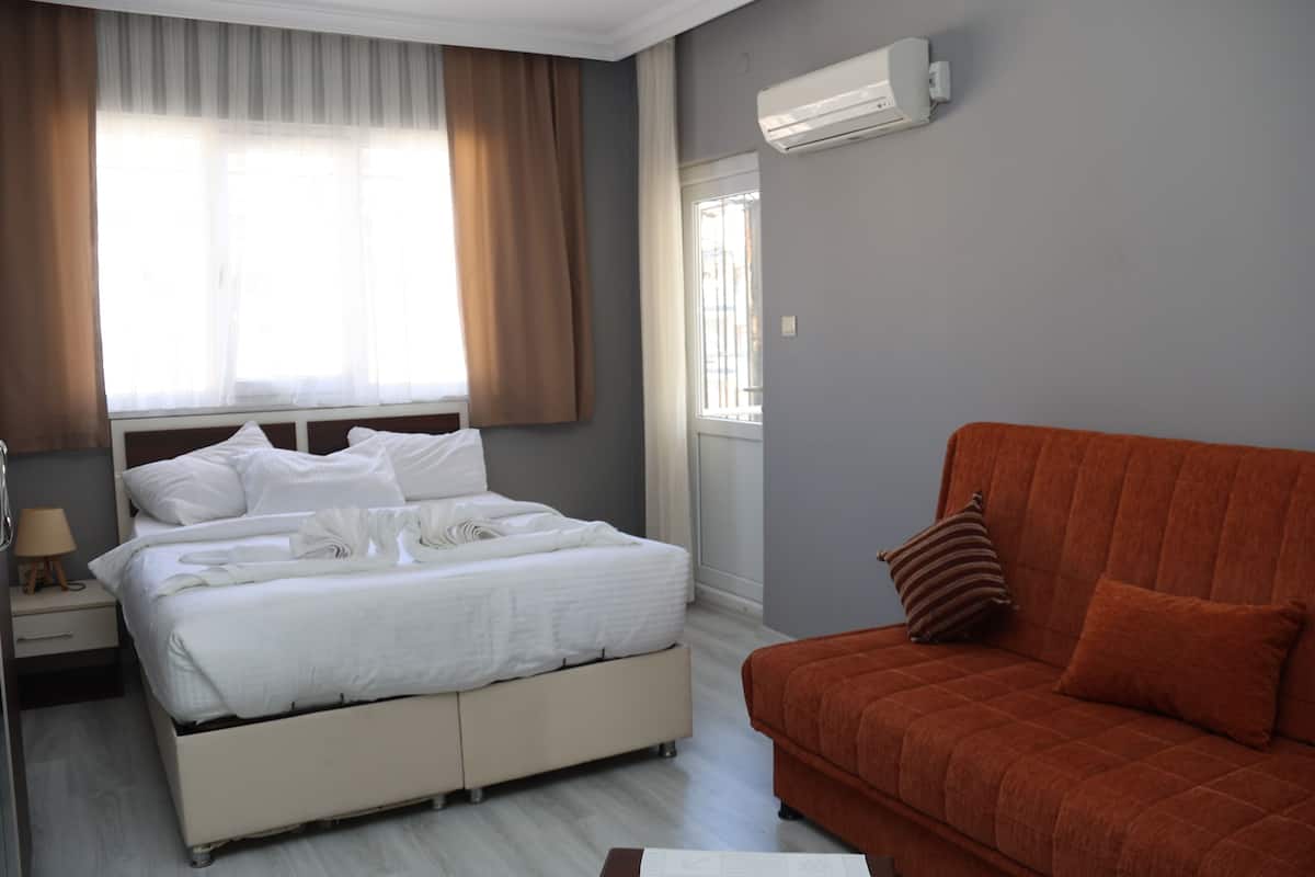 A bedroom with a bed, a nightstand, a lamp, a window with curtains, an air conditioner, and a sofa.