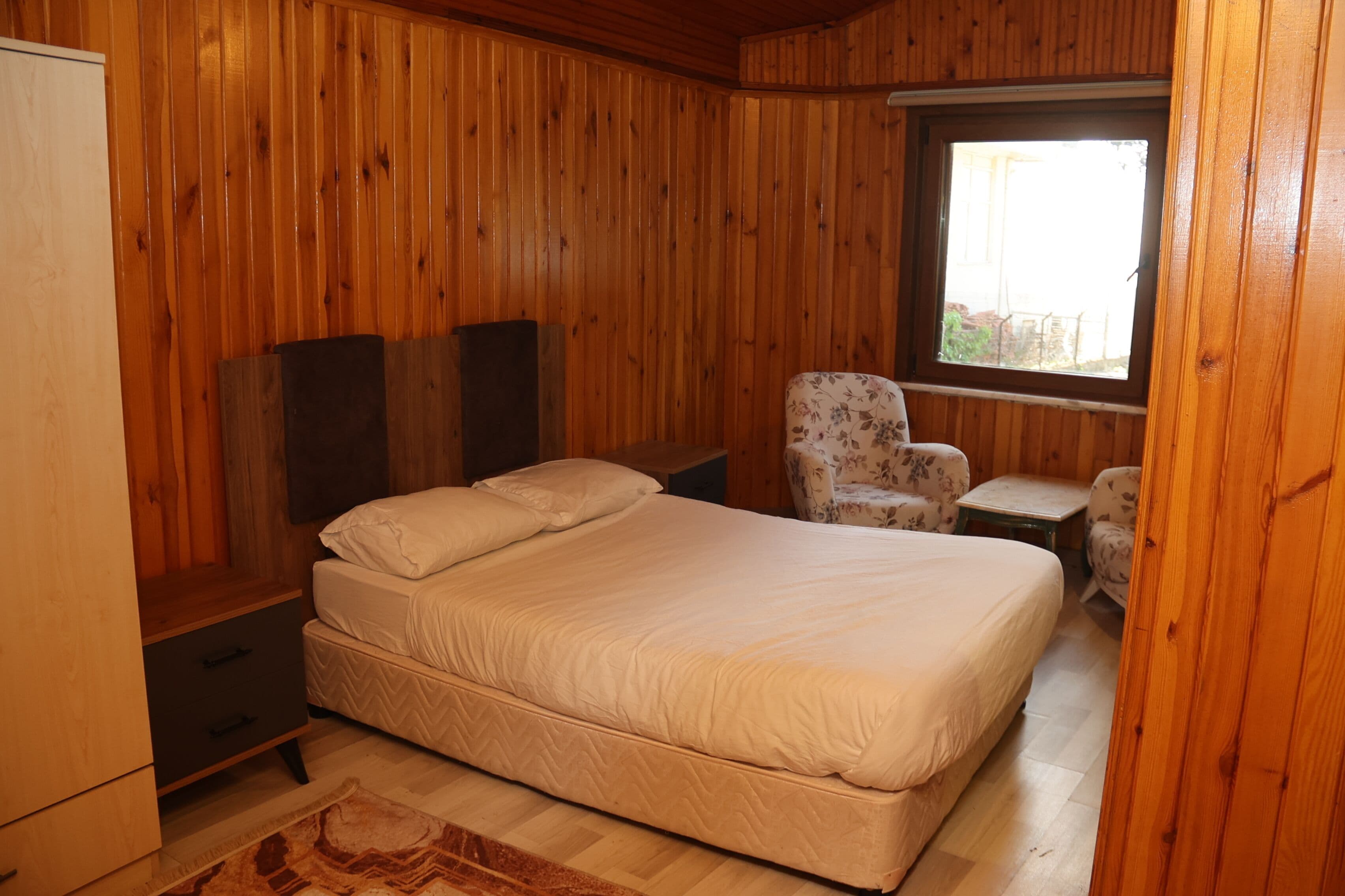 A wooden-paneled bedroom with a bed, two nightstands, a chair, and a small table.