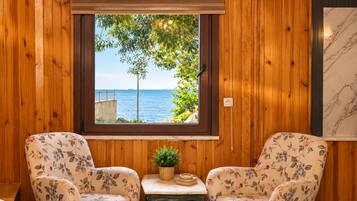 A room with wooden paneling, a window with a view of the sea, two floral armchairs, a small table with a plant, and a marble-framed artwork.