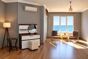Une pièce avec un plancher en bois, un miroir rond au-dessus d’une commode, une fenêtre donnant sur la mer et une table avec des chaises.