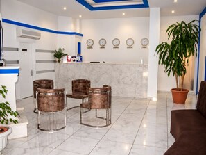 Vestíbulo de hotel con mostrador de recepción, cuatro sillones de cuero, una planta grande en maceta y relojes que muestran la hora de diferentes ciudades.