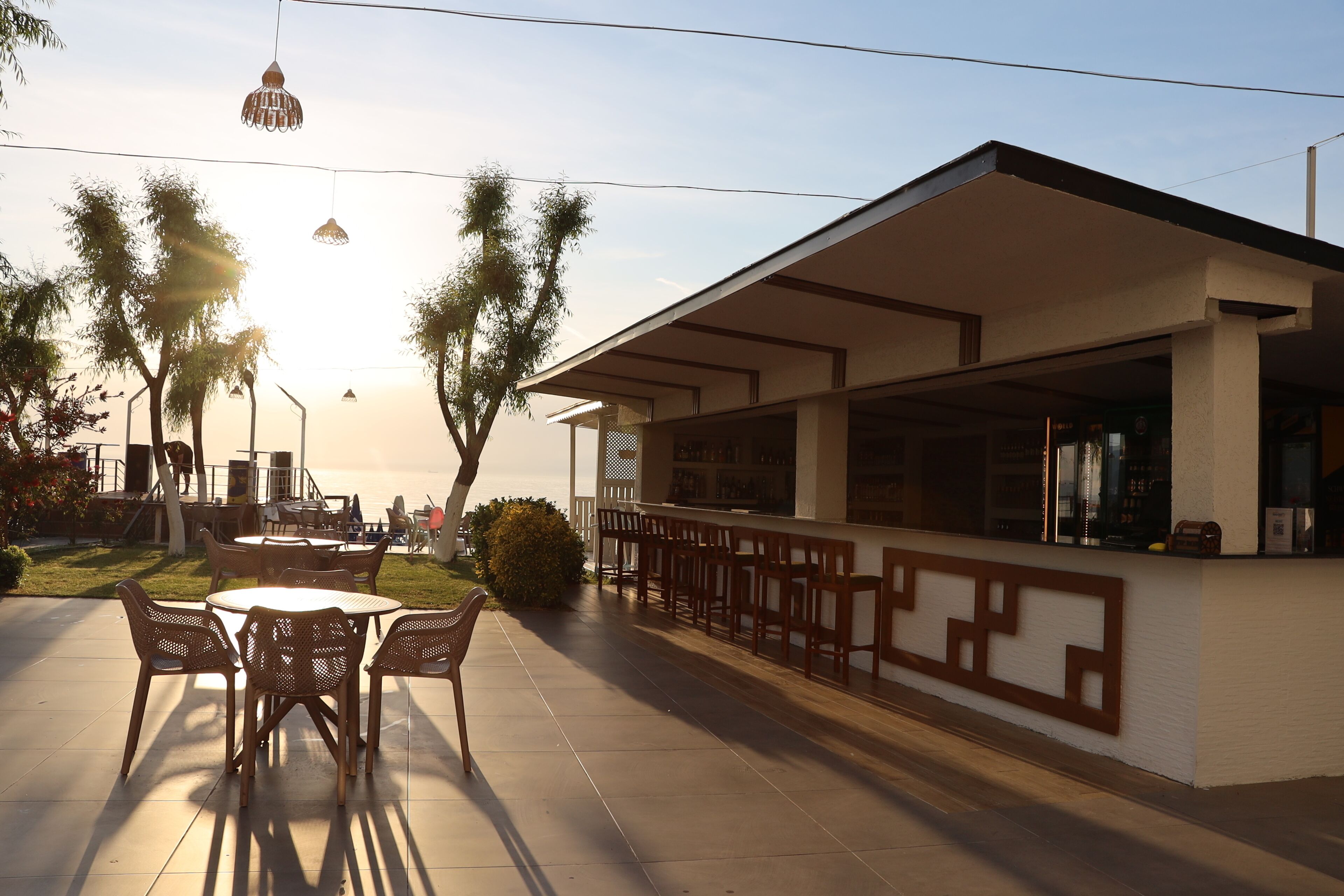 An outdoor seating area with tables and chairs, a bar with a white facade, and palm trees in the background.