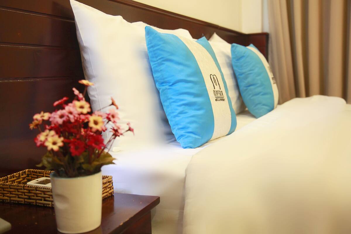 A hotel room with a neatly made bed, a bedside table with a vase of flowers, and a headboard with pillows.