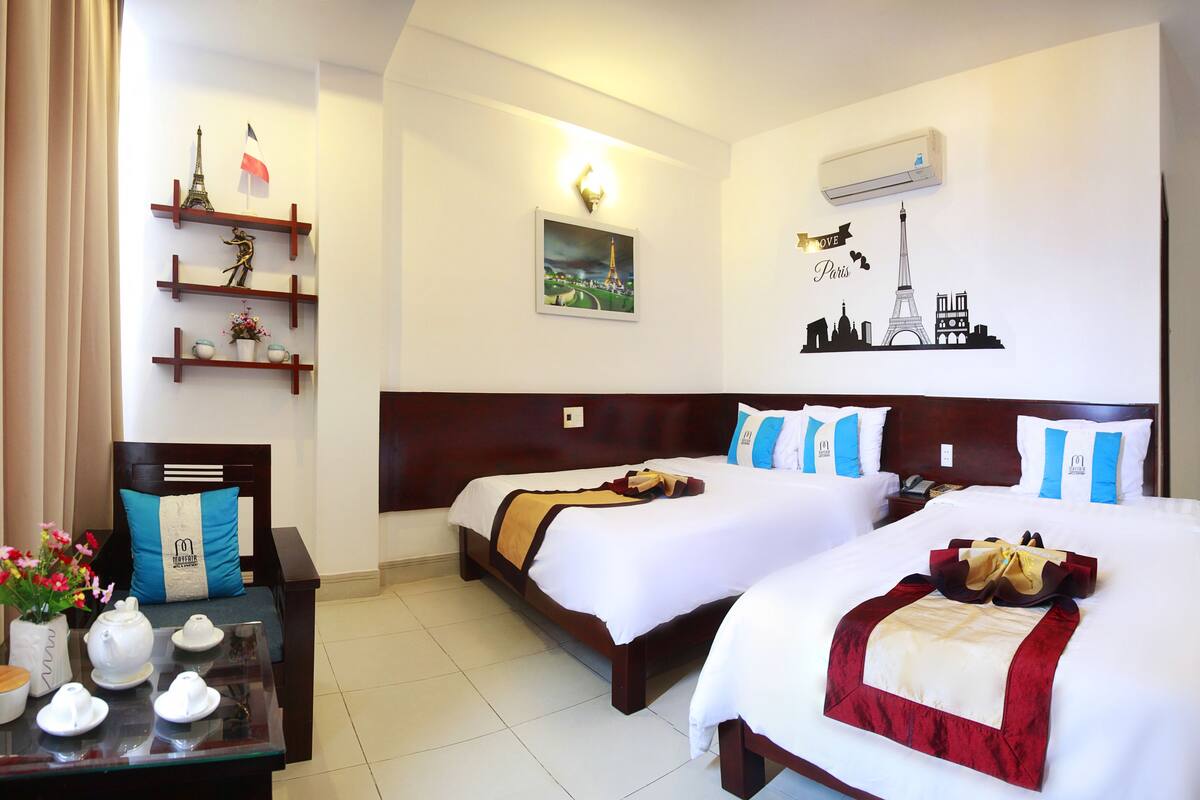 A hotel room with two beds, a nightstand, a table with tea sets, and wall decorations including the Eiffel Tower and a cityscape.