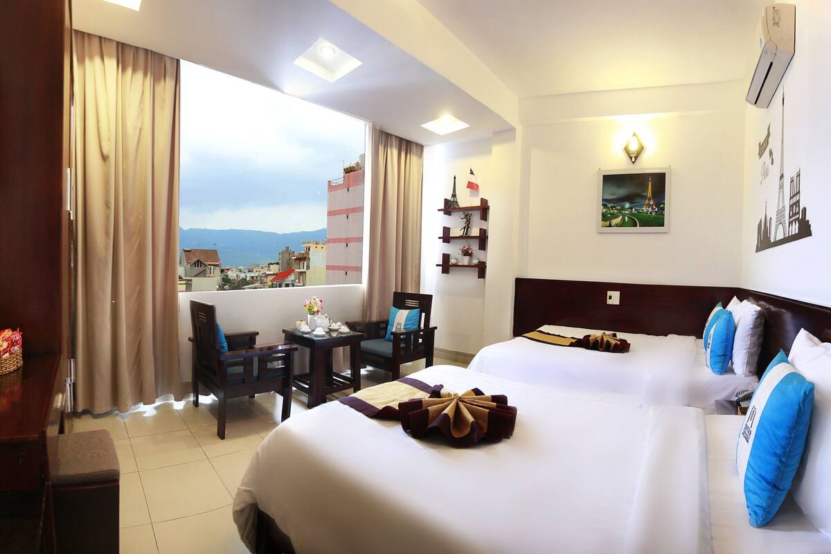 A hotel room with a bed, a desk, a chair, a small table with a vase of flowers, and a view of a cityscape through the window.