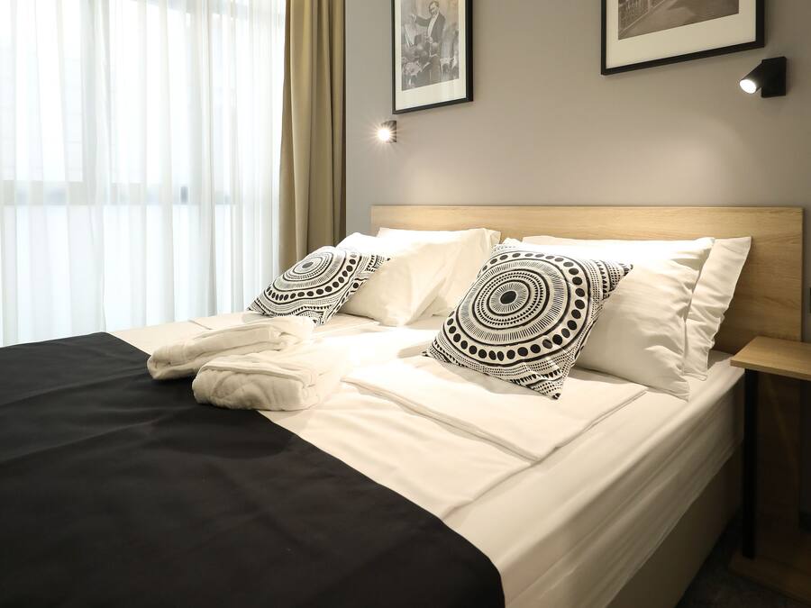 Een net opgemaakt bed met witte lakens en gestreepte kussens, een houten hoofdbord en twee ingelijste foto’s aan de muur.