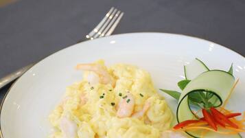 Un plato de huevos revueltos con camarones sobre un plato blanco, acompañado de una guarnición de pepino y zanahoria, sobre una mesa con un tenedor y un vaso de agua al fondo.