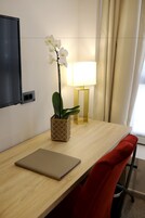 Habitación de hotel con un escritorio de madera, una laptop, una silla roja, una orquídea en maceta, una lámpara y un televisor montado en la pared.