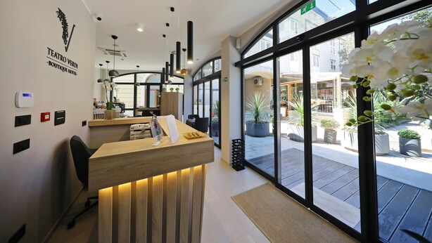 Un espace d’accueil d’hôtel avec un comptoir de réception en bois, une grande fenêtre donnant sur la rue et une enseigne indiquant le nom de l’hôtel.