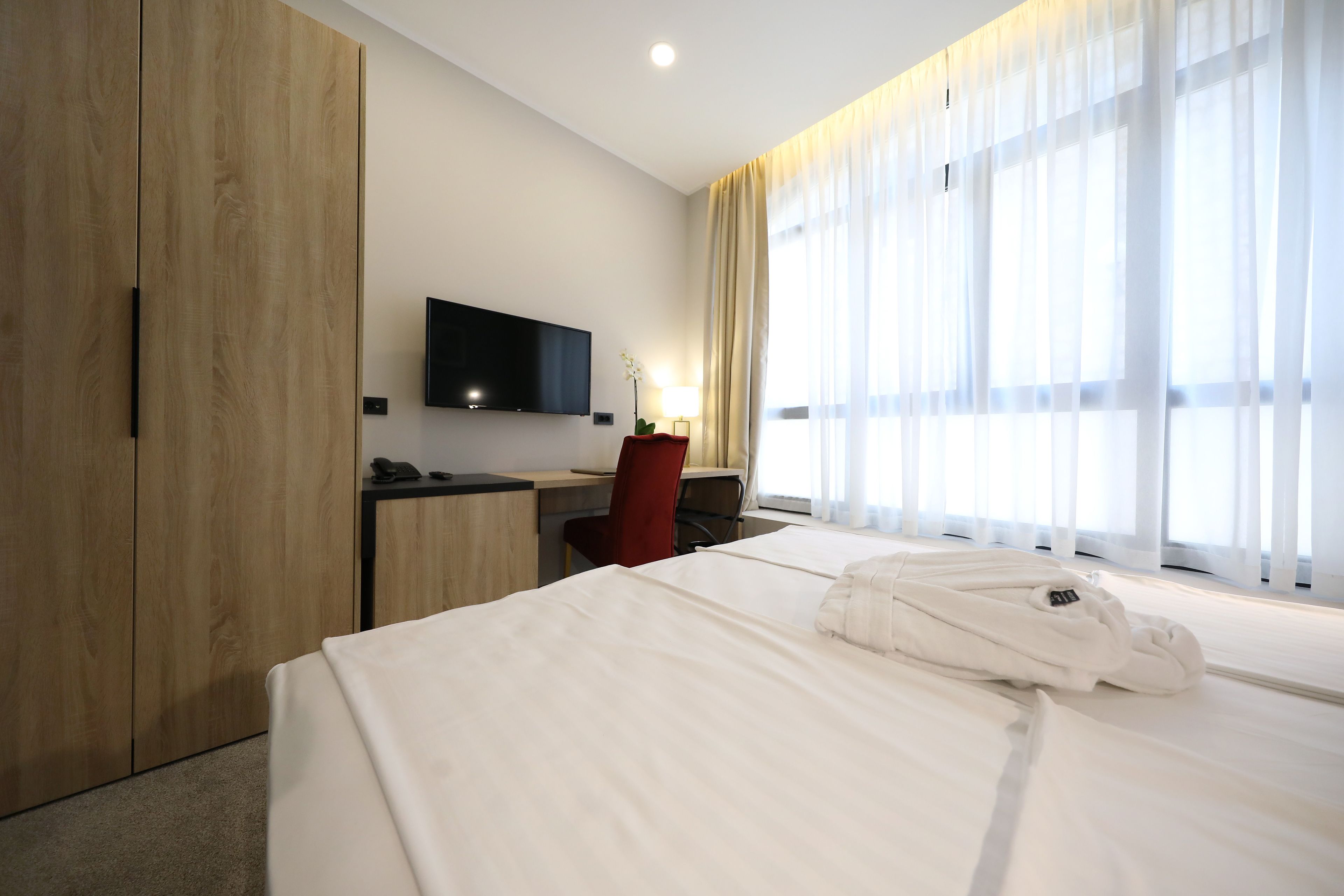 Habitación de hotel con una cama grande, un escritorio, una silla, un televisor montado en la pared y una ventana con cortinas translúcidas.