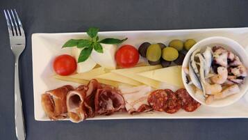 Un plato rectangular blanco con diversos alimentos, incluyendo tomates, aceitunas, queso y embutidos.
