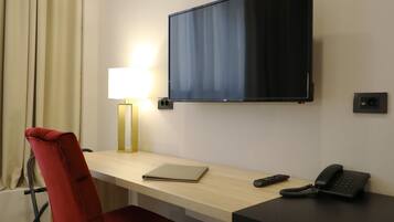 Quarto de hotel com escrivaninha, televisão, uma cadeira vermelha, abajur, telefone e cortina.
