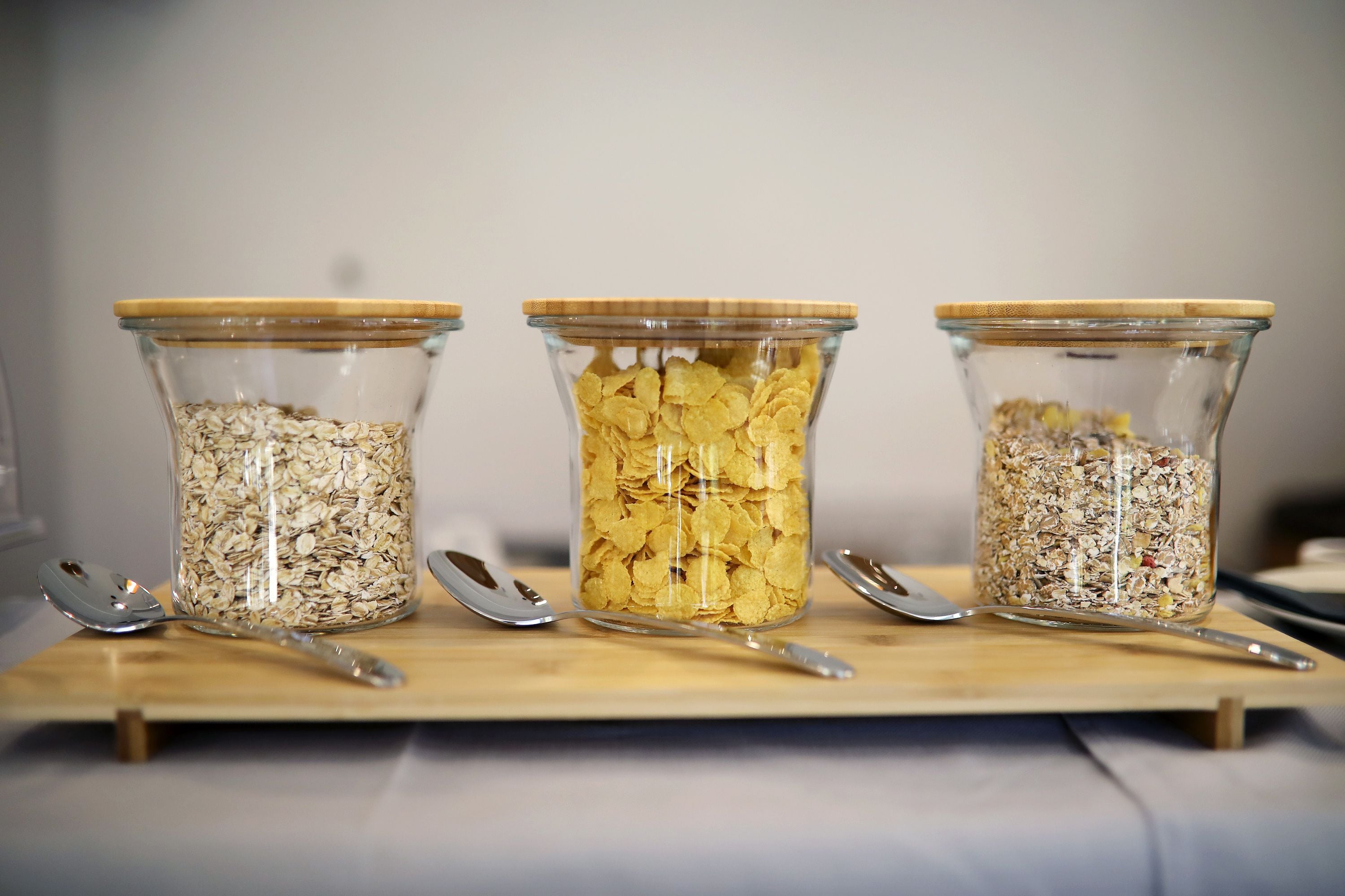 Three glass jars with wooden lids on a wooden tray, each containing different types of cereal.
