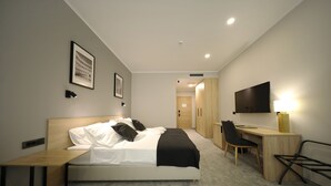 Une chambre d’hôtel moderne, équipée d’un lit, d’une table de chevet, d’un bureau, d’une chaise, d’une télévision et d’un tableau encadré au mur.