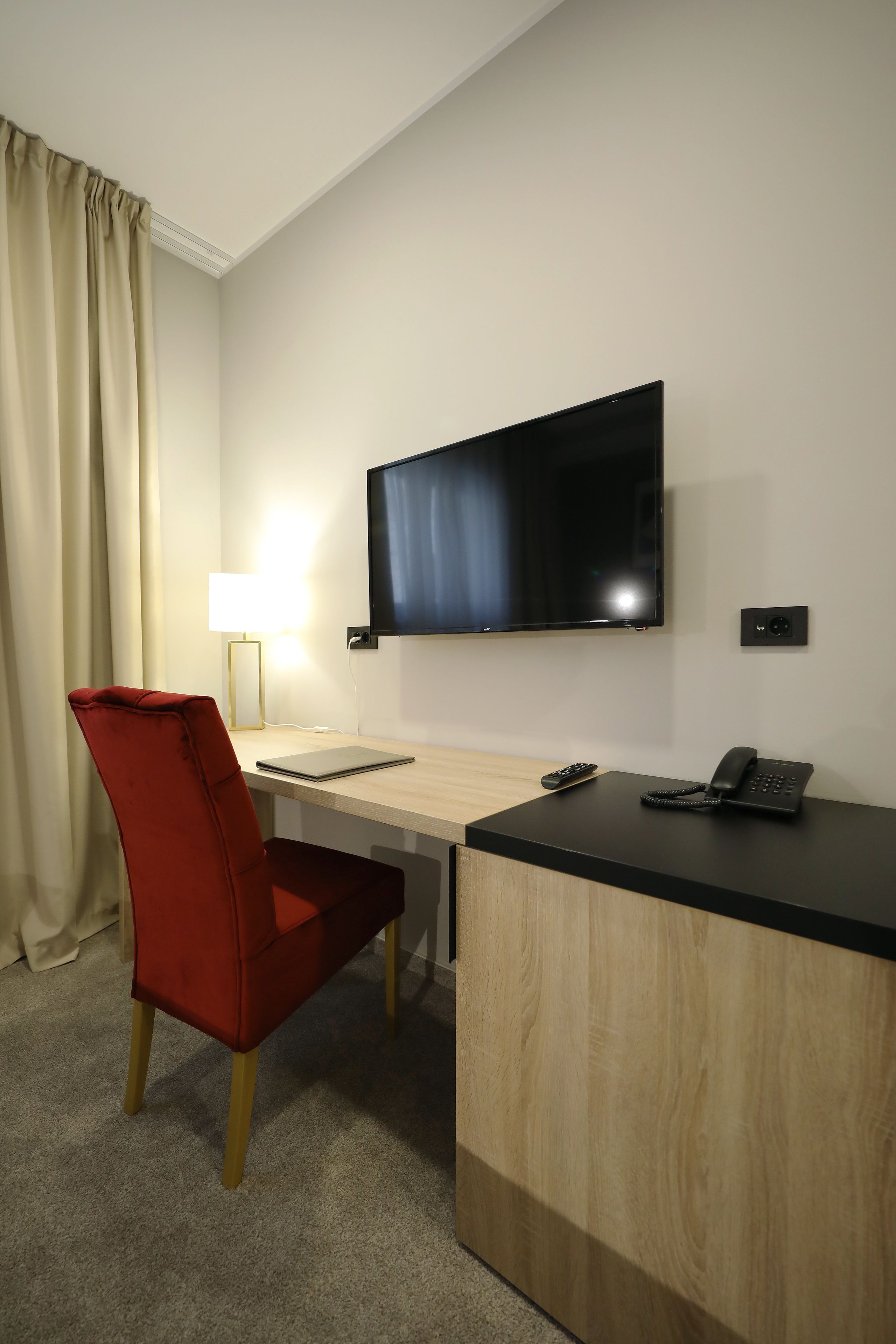 A hotel room with a flat-screen TV mounted on the wall, a desk with a lamp, a red chair, and a phone on the desk.