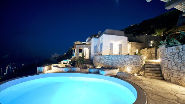 A swimming pool with a stone wall and steps leading to a white building at night. - Villa Ammonite (Mykonos)