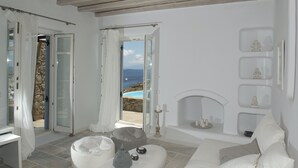 A modern, minimalist living room with a stone wall, a fireplace, and a view of the sea. - Villa Ammonite (Mykonos)