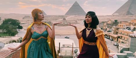 Dos personas con vestimenta histórica observan un paisaje urbano con pirámides al fondo.