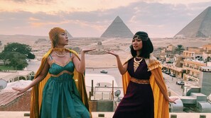 Two individuals in historical costumes overlooking a cityscape with pyramids in the background.