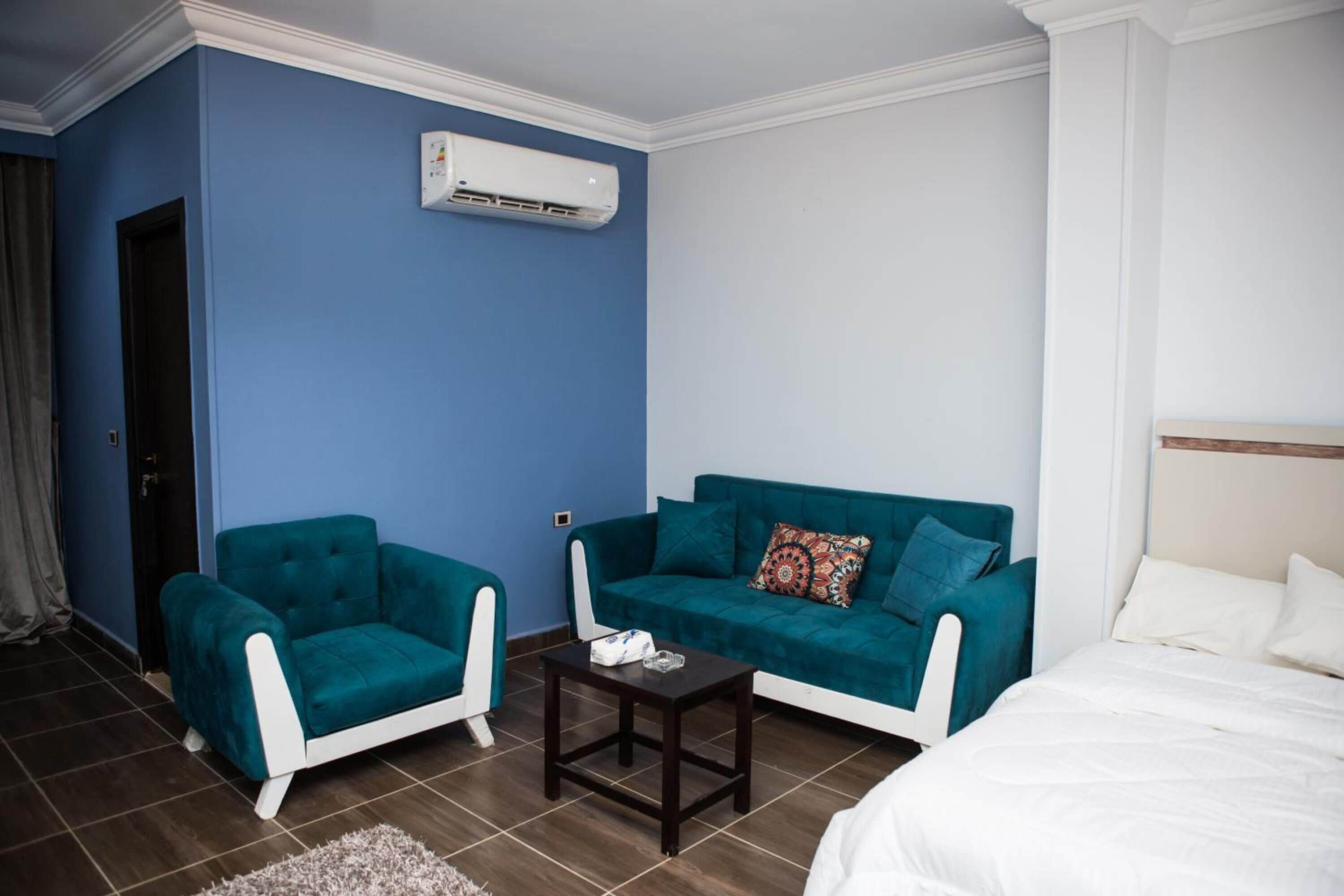 A hotel room with a blue wall, a teal sofa, a white armchair, a small wooden table, and a bed with white bedding.