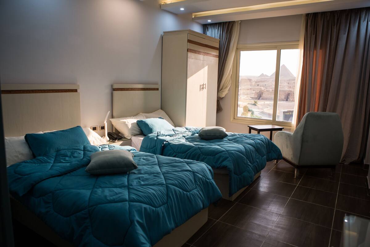 A hotel room with two beds, a wardrobe, a chair, and a window with a view of a pyramid.