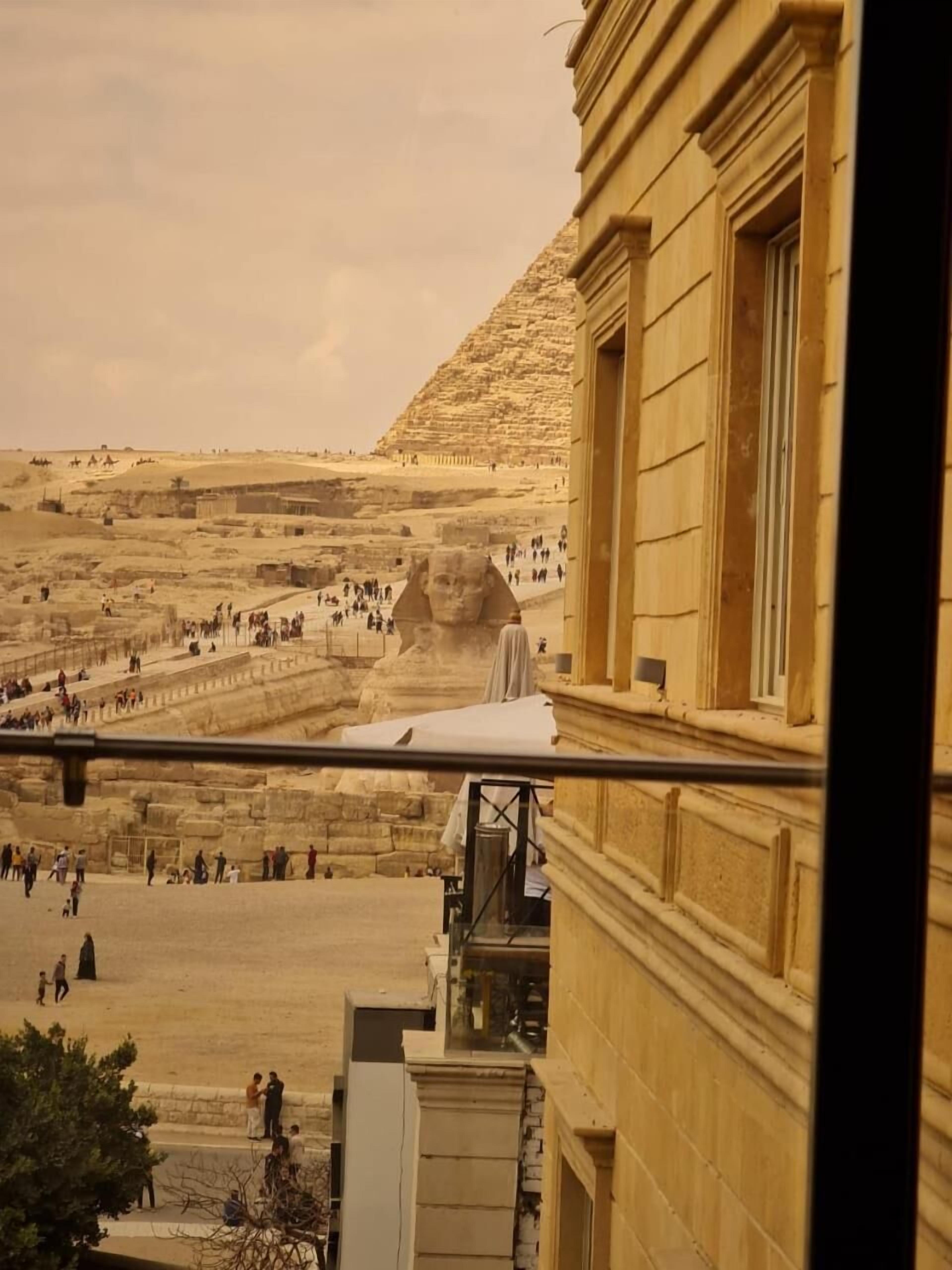 A view of the Sphinx with tourists in the background, a building with classical architecture, and a clear sky.