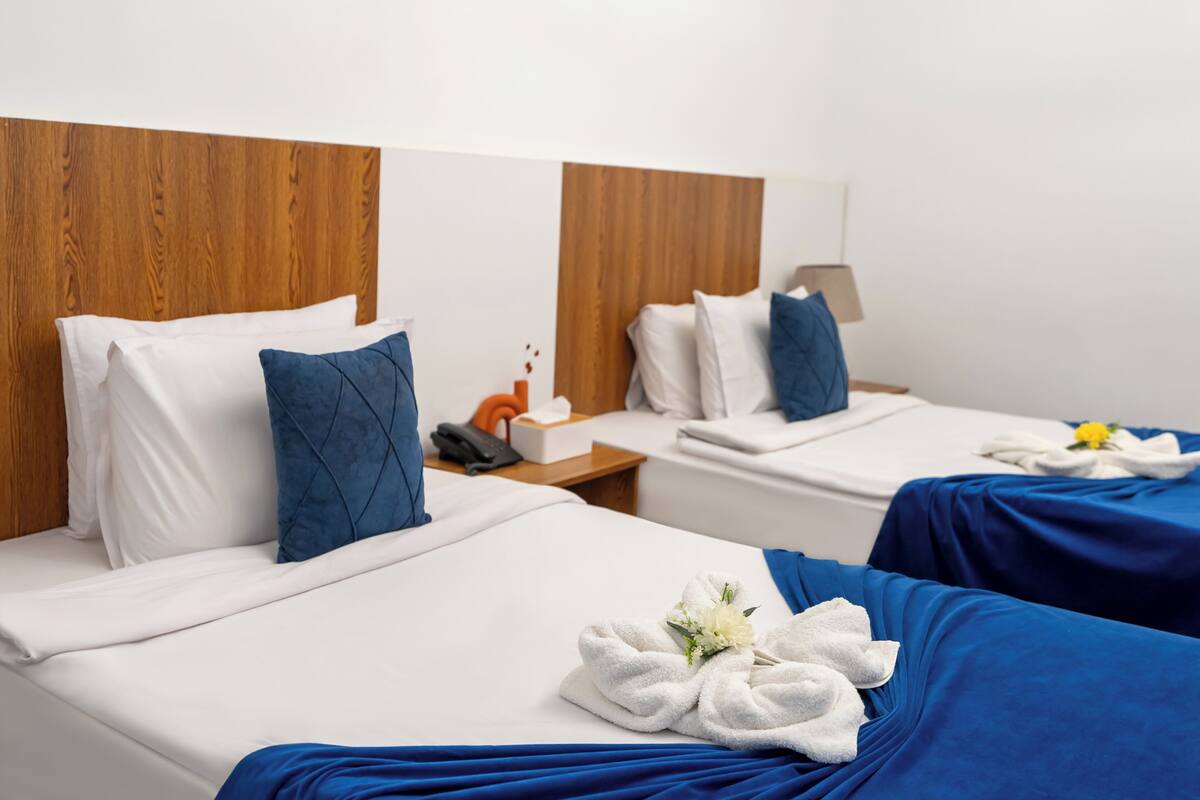 A hotel room with two beds, each with white linens and blue accents. There is a wooden headboard, a bedside table with a lamp, and a small table with a phone and tissues.