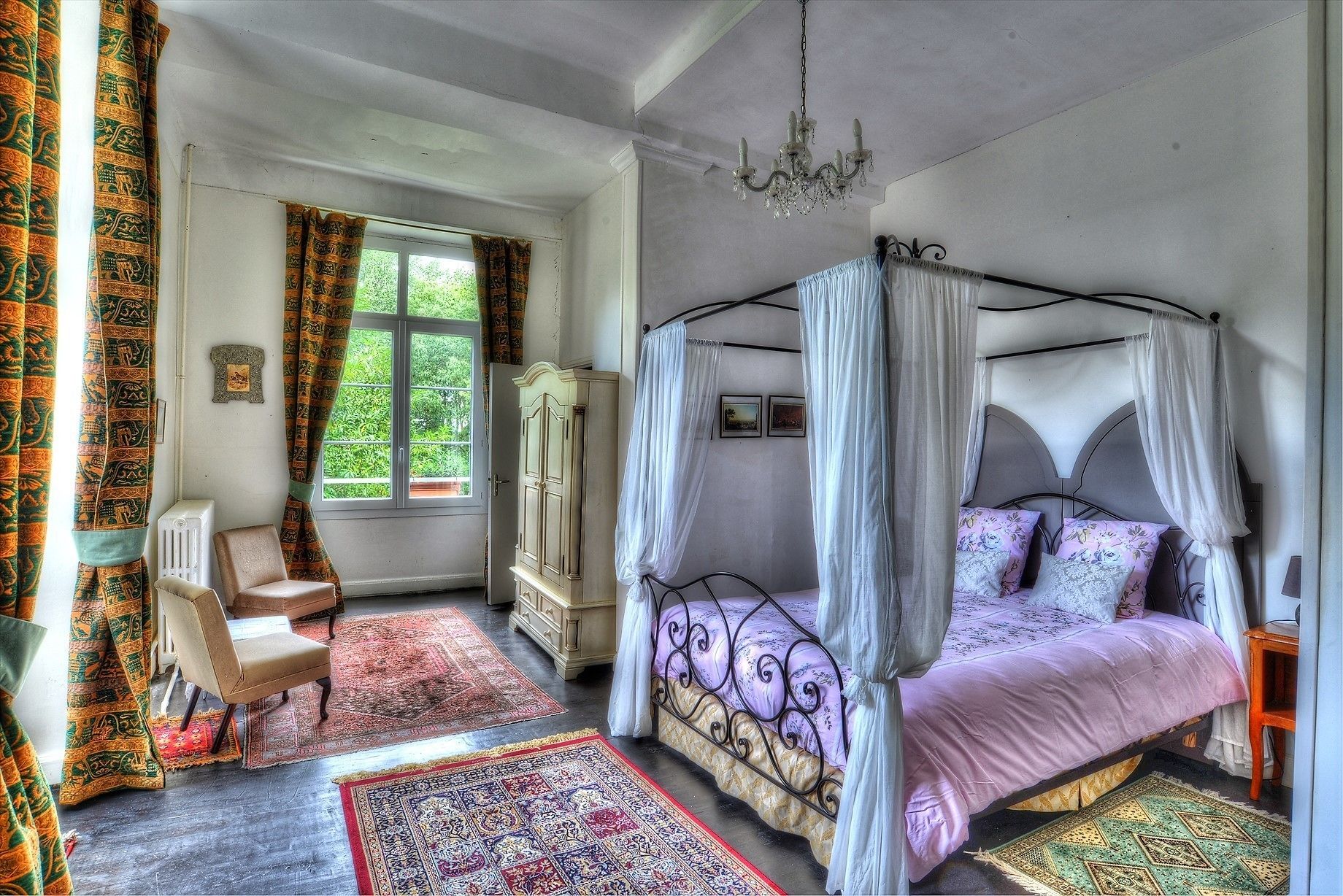 Een slaapkamer met een hemelbed, een zithoek met een stoel en een tafel, en een groot raam met gordijnen.
