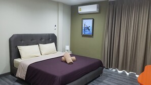 A hotel room with a large bed, a bedside table, an air conditioning unit, a framed picture on the wall, and a window with curtains.