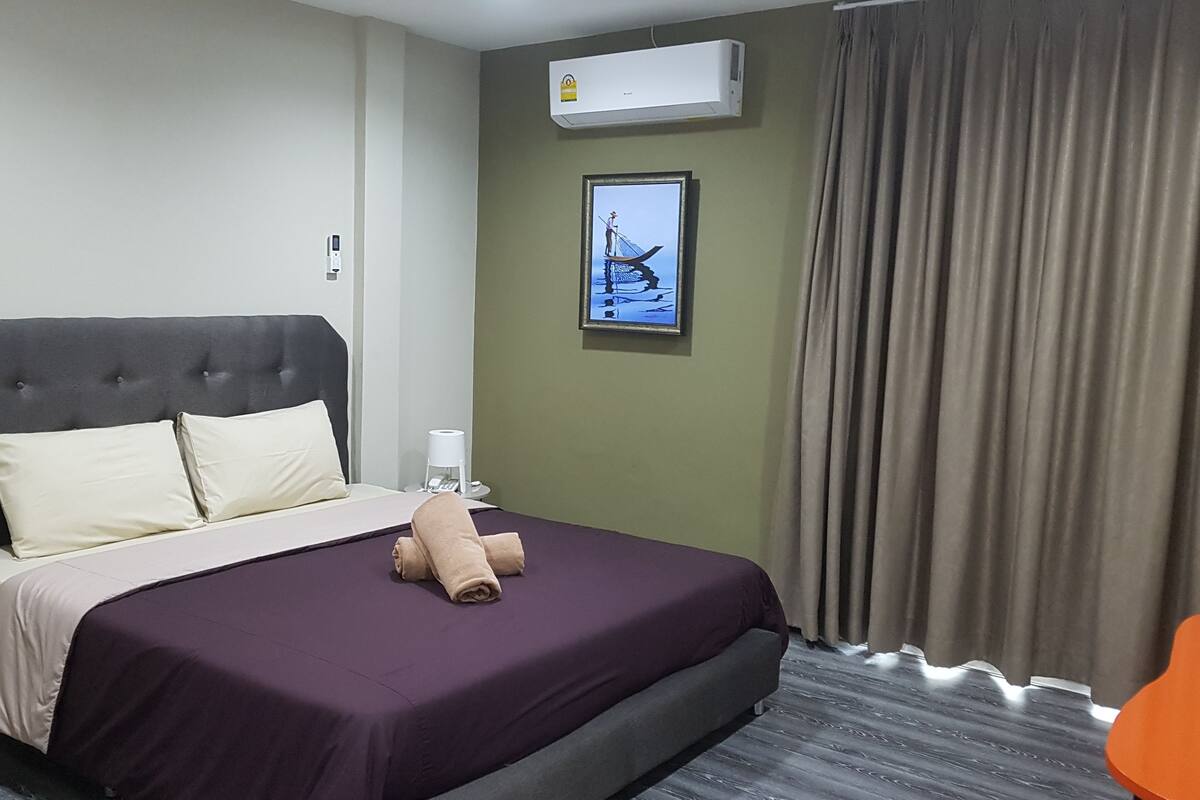 Een hotelkamer met een groot bed, een nachtkastje, een airconditioningapparaat, een ingelijst schilderij aan de muur en een raam met gordijnen.