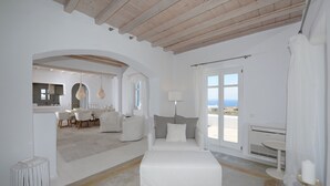 Una habitación amplia con una cama grande, un área de descanso con una mesa y sillas, y vista al mar a través de amplios ventanales.