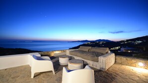 Un patio frente al mar con muebles de mimbre, un muro de piedra y vistas al océano y colinas lejanas.