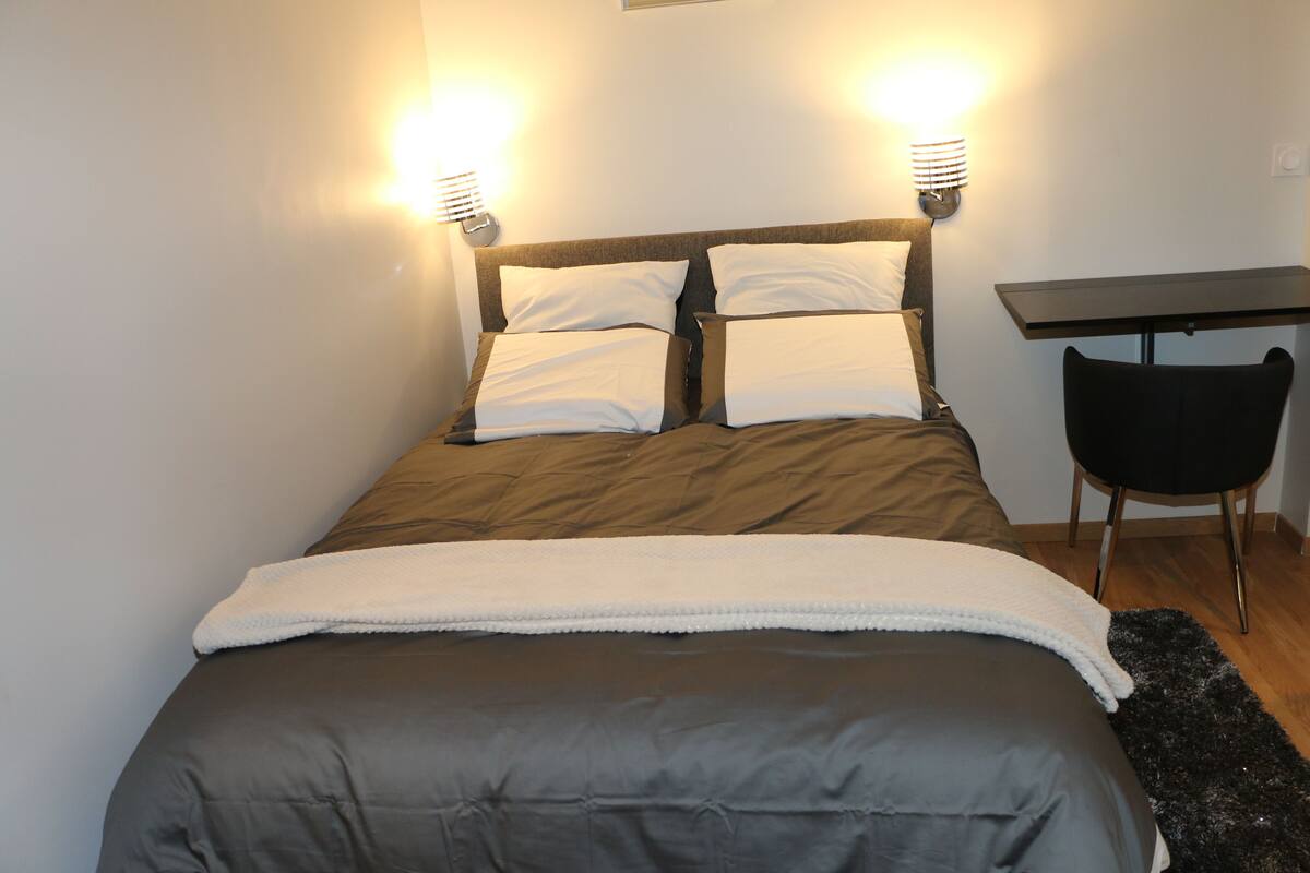 A neatly made bed with a dark-colored duvet and pillows, a small desk with a chair, and two wall-mounted lamps.