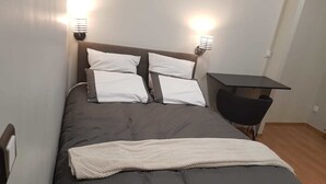 Uma cama com edredom cinza escuro, almofadas brancas e um cobertor branco. Há duas luminárias fixadas na parede, de cada lado da cama.