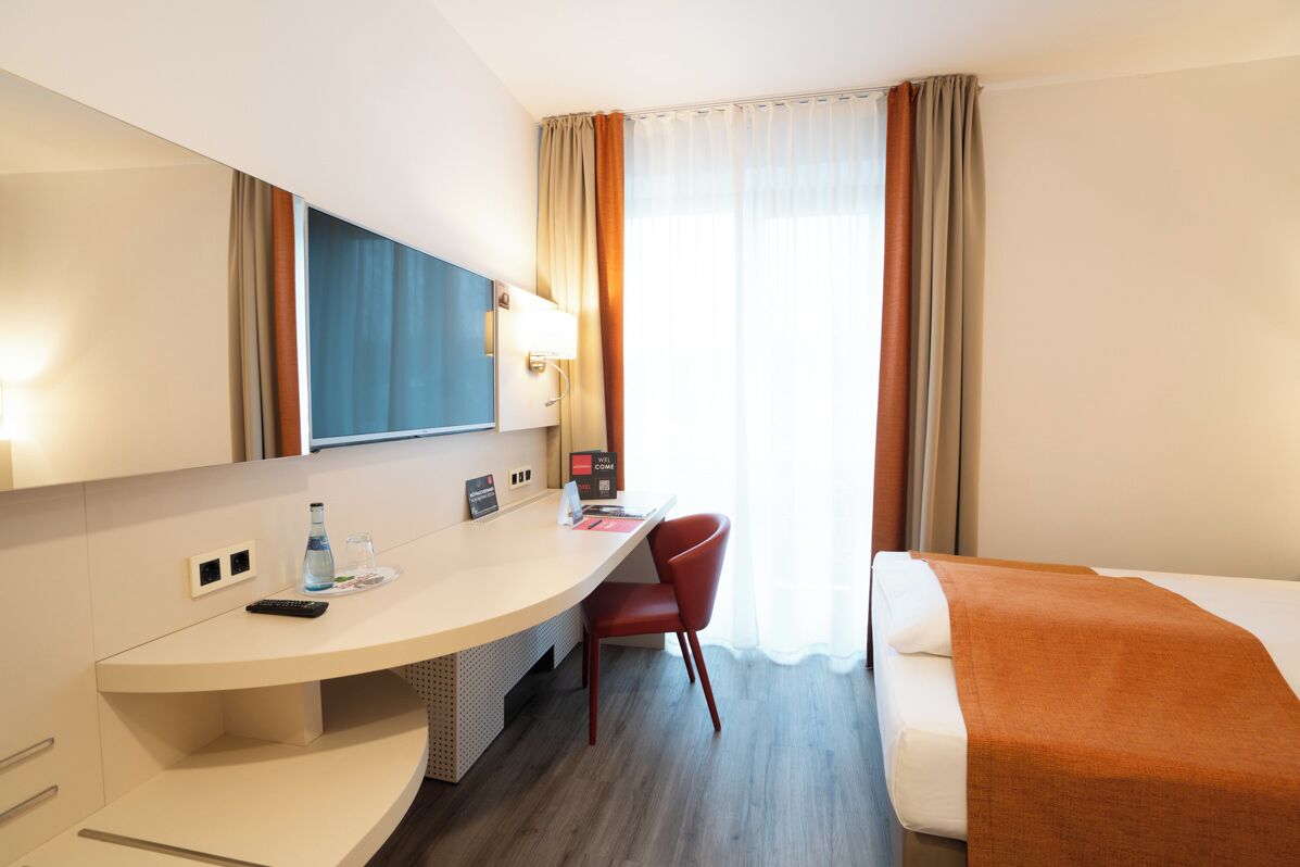 A hotel room with a bed, a desk with a chair, a TV, and a window with curtains.