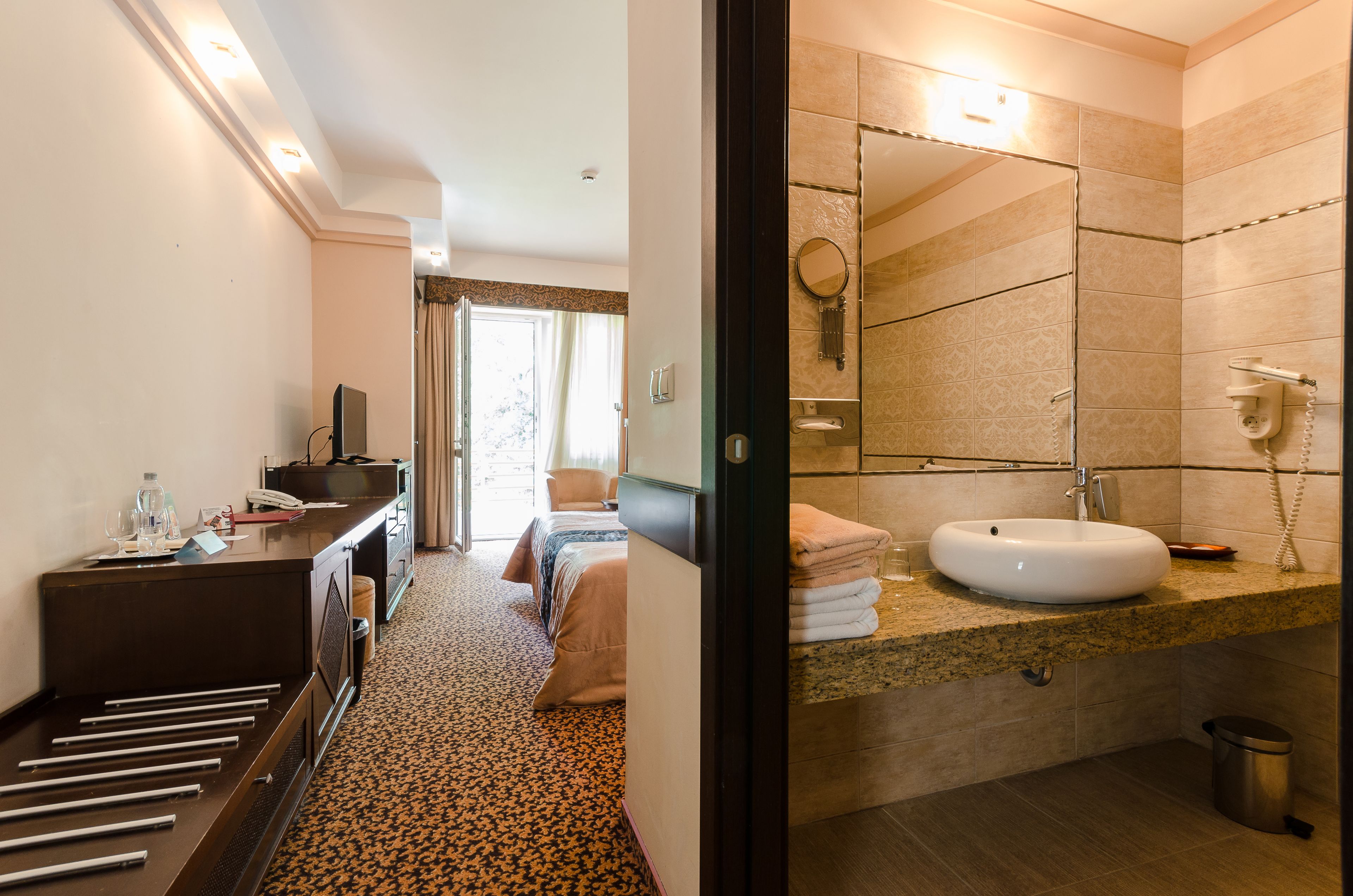 Quarto de hotel com cama, mesa de cabeceira e banheiro com pia, espelho e porta-toalhas.