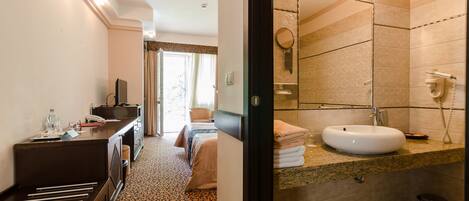 Habitación de hotel con cama, mesita de noche y baño con lavamanos, espejo y perchero para toallas.