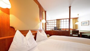 Habitación de hotel con dos camas, cabeceros de madera, una ventana con cortinas, una mesita pequeña y una zona de estar con una silla y un sofá.
